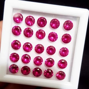 Pode incluir: Uma coleção de vinte e uma pedras preciosas redondas, facetadas, vermelho escuro, exibidas numa bandeja branca. As gemas estão uniformemente espaçadas e refletem a luz, mostrando o seu corte brilhante e a sua cor rica.