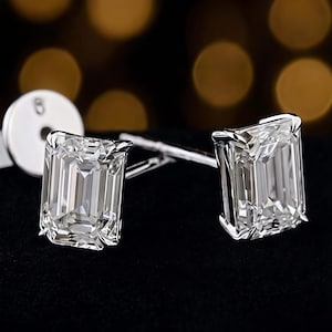 May include: A pair of emerald-cut stud earrings featuring clear, faceted stones set in polished silver-toned metal. Each earring has four prongs securing the rectangular stone, with a circular earring back visible behind one of the studs.