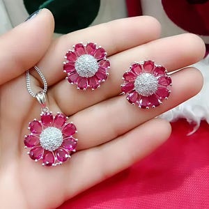 May include: A jewelry set featuring a pendant necklace and matching stud earrings. Each piece is designed as a flower with ruby-red petals and a central cluster of sparkling white stones. The set is displayed on a pink surface.