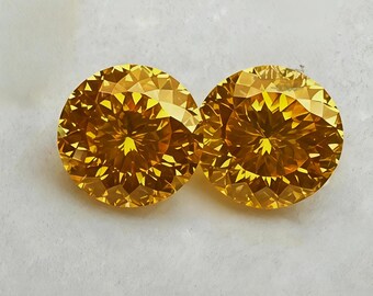 May include: Two round-cut gemstones with a vibrant golden-yellow hue. Each stone features a faceted surface with a symmetrical pattern of triangular and kite-shaped cuts that reflect light, creating a brilliant, sparkling appearance.