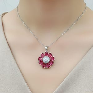 May include: A silver necklace with a flower pendant. The pendant features a central cluster of small, white gemstones surrounded by deep red gemstone petals. The necklace is set against a neutral background.
