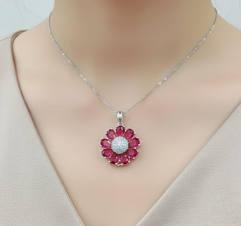 May include: A silver necklace with a flower pendant. The pendant features a central cluster of small, white gemstones surrounded by deep red gemstone petals. The necklace is set against a neutral background.