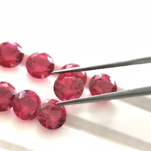 May include: A collection of faceted, round, deep red gemstones. One gemstone is being held by a pair of silver tweezers. The gems are arranged on a white surface, showcasing their brilliant cut and color.