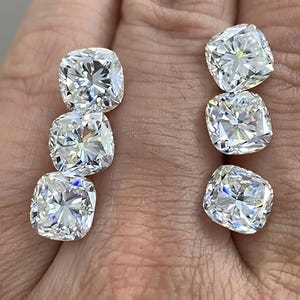 以下が含まれることがあります： 透明で無色、高い輝きを放つ5つのクッションカットのルースストーン。それぞれの石は光を反射するファセットカットが施されており、肌色の表面に2列の縦並びで配置され、その透明度の高さを示しています。