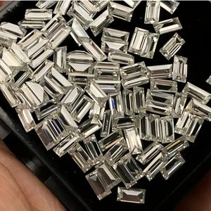 Puede incluir: Una colección de gemas claras, de corte rectangular, posiblemente diamantes, se dispersan sobre una superficie negra. Las gemas son muy reflectantes, captando la luz y creando un efecto brillante.