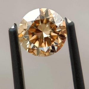 May include: A round, faceted diamond with a warm, champagne color. The gemstone is held by black tweezers, showcasing its brilliance and clarity. The diamond's facets reflect light, creating a sparkling effect.