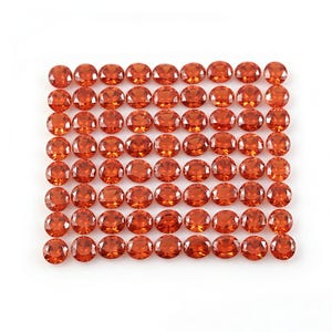 Puede incluir: Una colección de gemas de color naranja, talladas en forma ovalada, dispuestas en un patrón de cuadrícula. Las gemas tienen una superficie facetada que refleja la luz. La disposición es simétrica, con gemas de tamaño y forma similares. Fondo blanco.