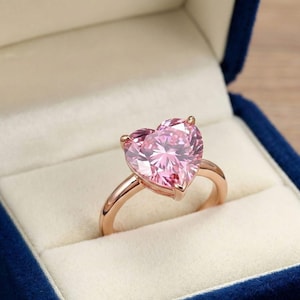 Op de afbeelding: Een roségouden ring met een grote, hartvormige roze edelsteen. De ring wordt gepresenteerd in een blauwe fluwelen ringdoos met een witte satijnen binnenkant. De ring heeft een warme, metallic kleur.