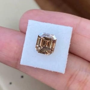 May include: A rectangular, light brown gemstone with a step cut, resting on a white square. The gemstone has a reflective surface and a complex internal structure, showcasing its facets. The overall appearance is elegant and luxurious.