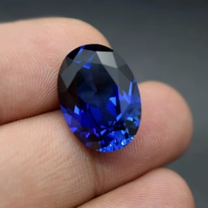 May include: A deep blue, oval-cut gemstone. The facets catch the light, creating a sparkling effect. The gem is held in a hand, highlighting its colour and shape. The stone's dimensions are not visible.