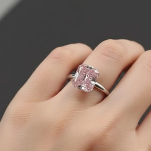 May include: A radiant-cut pink diamond ring set on a silver band. The large, rectangular gemstone is a soft pink hue, catching the light. The ring is displayed on a hand against a dark background.