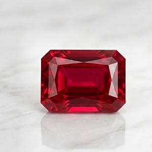May include: A vibrant, deep red gemstone cut in a rectangular emerald shape with faceted surfaces that create sharp geometric reflections. The stone displays a rich, saturated crimson hue throughout its transparent body, showcasing precise symmetry.
