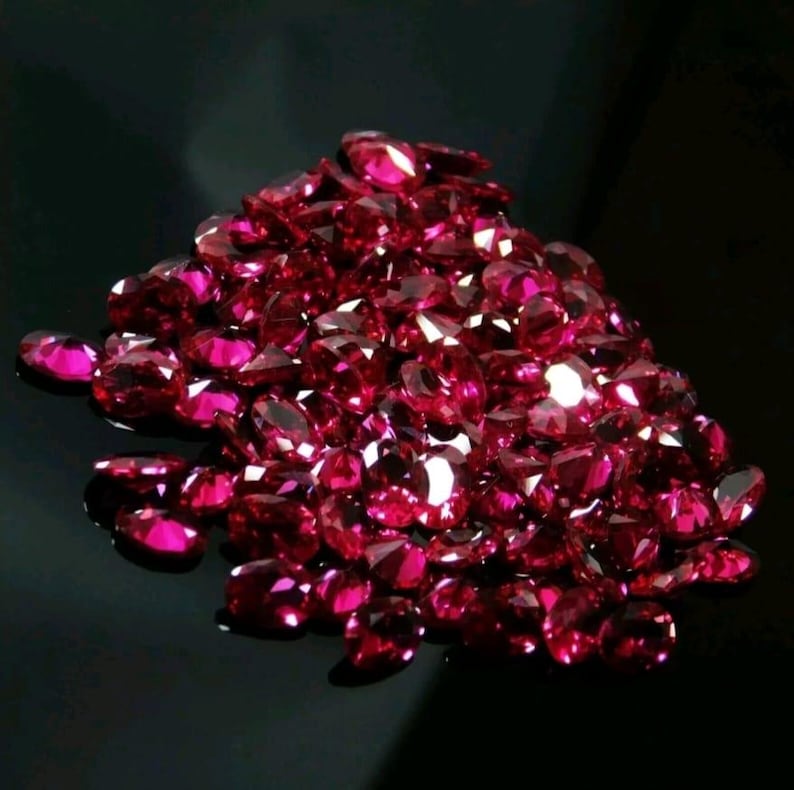 May include: A pile of faceted, oval-cut, deep red gemstones. The gems are clustered together, reflecting light and creating a sparkling effect against a dark background. The gemstones are likely rubies.
