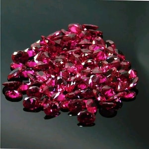 May include: A pile of faceted, deep red gemstones. The gems are scattered across a reflective black surface, catching the light and displaying various shapes and sizes. The overall effect is a display of rich color and sparkle.