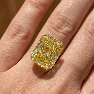 Könnte beinhalten: Ein großer, rechteckig geschliffener gelber Diamantring. Der Edelstein hat eine leuchtende, goldene Farbe und ist auf einem schlichten Band gefasst. Der Ring wird an einem Finger gezeigt, fängt das Licht ein und zeigt seine Brillanz.