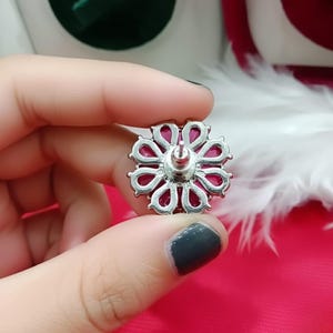 May include: A silver-coloured floral stud earring with a vibrant magenta centre. The earring features a detailed, openwork design with a post for secure wear. The earring is held in a hand against a pink background.