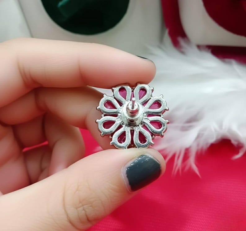 May include: A silver-coloured floral stud earring with a vibrant magenta centre. The earring features a detailed, openwork design with a post for secure wear. The earring is held in a hand against a pink background.