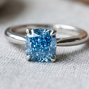 Op de afbeelding: Een zilveren ring met een grote, vierkant geslepen, lichtblauwe edelsteen. De edelsteen is gezet met vier klauwen en heeft vele facetten. De ringband is glad en gepolijst. De achtergrond is een lichtgrijze stof.