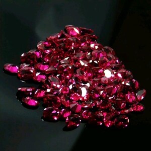 May include: A pile of faceted, oval-cut, deep red gemstones. The gems are clustered together, reflecting light and creating a sparkling effect against a dark background. The gemstones are likely rubies.