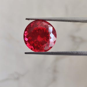 May include: A faceted, round, bright red gemstone is held by a pair of silver tweezers. The gem has a brilliant cut, reflecting light. The background is a soft, neutral color. This image showcases a gemstone, likely for jewelry or collection.