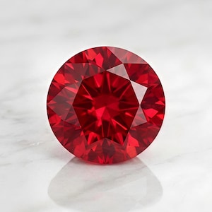 May include: A round, brilliant-cut gemstone with a deep, vibrant red color. The stone features numerous triangular and kite-shaped facets that create a complex, symmetrical pattern of light reflection and refraction across its circular surface.