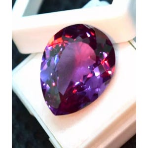 May include: A large, pear-shaped gemstone in shades of purple, with flashes of red and pink. The cut facets catch the light, creating a sparkling effect. The gem is presented against a white background.