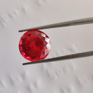 Könnte beinhalten: Ein runder, facettierter, leuchtend roter Edelstein, der von einer Metallpinzette gehalten wird. Der Stein hat einen Brillantschliff mit zahlreichen dreieckigen Facetten, die das Licht reflektieren und eine tiefe, gesättigte karmesinrote Farbe erzeugen.