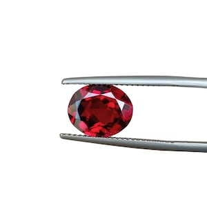 May include: A faceted, oval-shaped, deep red gemstone is held by a pair of silver tweezers against a white background. The gem's facets reflect light, creating a sparkling effect. The tweezers are positioned to hold the gem securely.