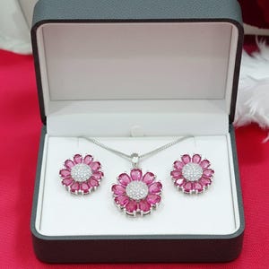 May include: A jewellery set comprising a necklace and earrings, displayed in a grey box. The set features a pendant and earrings, each shaped like a flower with pink petals and a central cluster of white stones. The jewellery is set against a white background.