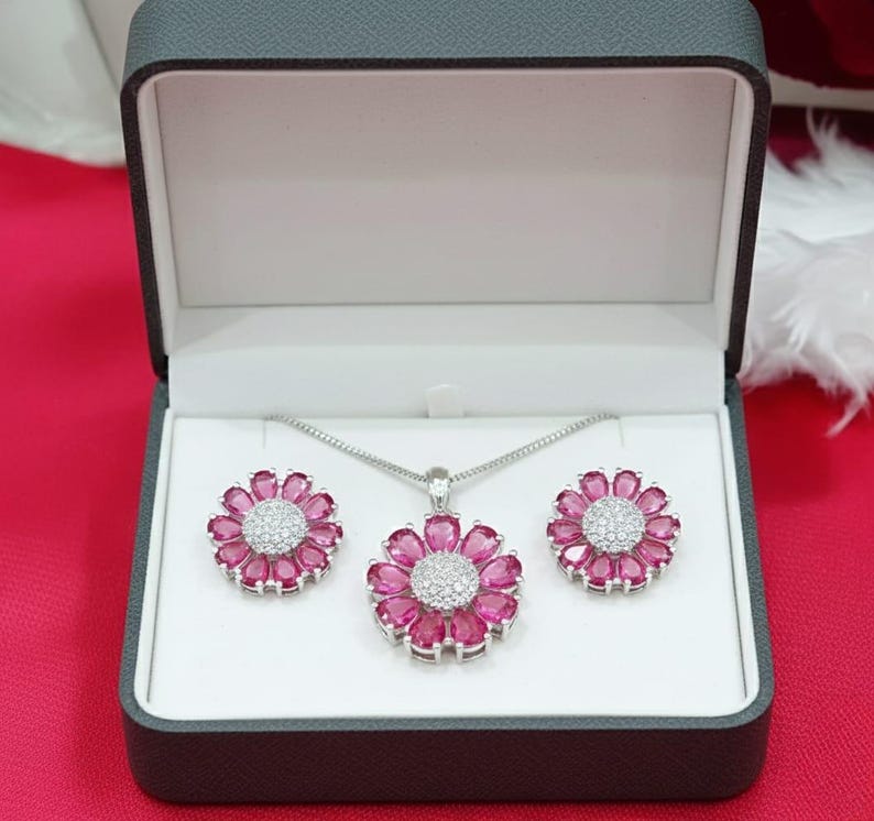 May include: A jewellery set comprising a necklace and earrings, displayed in a grey box. The set features a pendant and earrings, each shaped like a flower with pink petals and a central cluster of white stones. The jewellery is set against a white background.