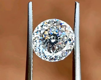 May include: A round, brilliant-cut diamond held by silver tweezers. The diamond sparkles with numerous facets, reflecting light. The background is a warm, neutral brown.