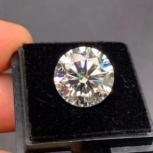 Puede incluir: Un diamante grande, redondo, con talla brillante y múltiples facetas, exhibido en un estuche negro forrado de terciopelo. El diamante brilla con reflejos internos, mostrando su claridad y talla. El estuche es sostenido por una persona.
