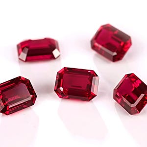 May include: Five faceted, rectangular-cut, deep red gemstones are displayed against a white background. The gems have a high degree of clarity and brilliance, reflecting light. The gems are likely rubies, and are a rich, saturated red color.