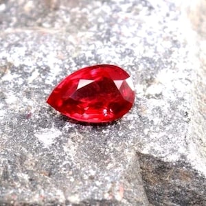 May include: A faceted, pear-shaped, red gemstone. The gem is a deep, vibrant red color with multiple facets that reflect light. The gemstone is set against a textured, gray stone surface.