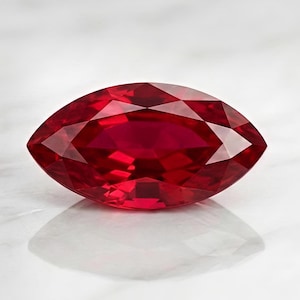May include: A marquise-cut ruby gemstone, displaying a deep red hue. The gemstone's facets reflect light, creating a brilliant sparkle. The ruby is set against a white background, highlighting its color and shape.