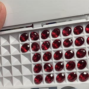 May include: A collection of oval-cut, deep red gemstones displayed in a white tray. The gems are arranged in rows, showcasing their polished surfaces and vibrant color. The tray is open, revealing the gemstones.
