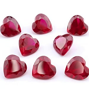 May include: Eight heart-shaped, faceted gemstones in a deep red hue are arranged on a white surface. The gems have a brilliant cut, reflecting light and creating a sparkling effect. The overall theme is jewelry and gemstones.