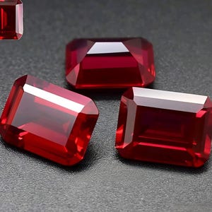 May include: Three deep red, rectangular, emerald-cut gemstones with faceted surfaces. The stones exhibit a high-gloss, transparent finish with sharp, clean edges and reflective highlights on their top facets, showcasing a rich, saturated crimson color.