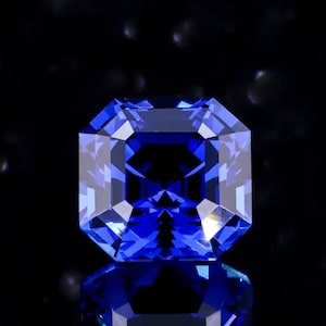May include: A faceted, square-cut, brilliant blue gemstone with a reflective surface. The gem is set against a dark background, showcasing its facets and internal structure. The gem's color is a deep, saturated blue.