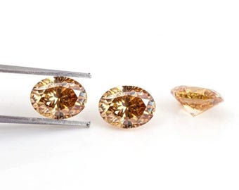 May include: Three oval-cut, light brown gemstones are displayed against a white background. One is held by tweezers, while the other two are positioned separately. The gems have a brilliant cut, reflecting light.