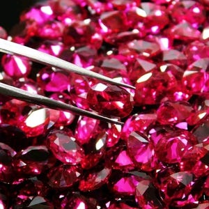 May include: Close-up of a collection of faceted, oval-cut, deep red gemstones. A pair of silver tweezers holds one gem, highlighting its brilliance. The gems are scattered, creating a rich, textured background.