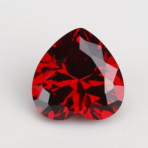 May include: A heart-shaped, faceted gemstone in a deep red hue. The gem's multiple surfaces reflect light, producing a sparkling effect. The symmetrical shape features a pointed base and a rounded top, suitable for jewellery.