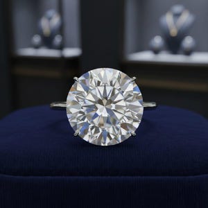 Puede incluir: Anillo de plata con un gran diamante de talla redonda. El diamante está sujeto por cuatro garras y descansa sobre una superficie de terciopelo azul oscuro. El fondo está borroso, lo que sugiere una joyería.