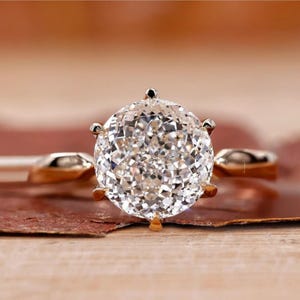 May include: A close-up of a gold ring featuring a large, round, clear diamond. The diamond is faceted, catching the light and creating a sparkling effect. The ring's band is a simple, polished gold. The ring is set against a neutral background.