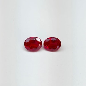Puede incluir: Dos gemas facetadas de corte ovalado, de color rojo intenso. Las gemas se muestran sobre un fondo blanco, resaltando su brillo y claridad. Las piedras son de tamaño y forma similares, lo que sugiere que son un par a juego.