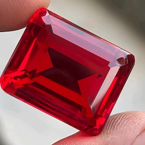 May include: A vibrant, transparent red gemstone with a rectangular emerald cut. The stone features sharp, clean facets that create a brilliant, reflective surface, showcasing a deep, saturated crimson hue throughout its entire crystalline structure.