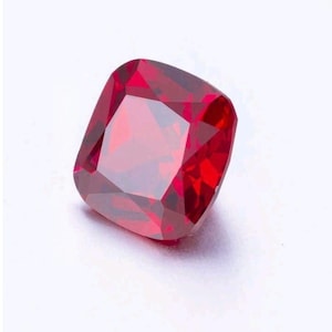 May include: A faceted, cushion-cut ruby gemstone. The gem is a deep, saturated red color and has a high degree of clarity. The facets create a brilliant display of light and shadow. The gemstone is set against a white background.