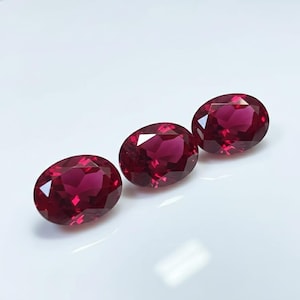 Op de afbeelding: Drie ovaalvormige, dieprode edelstenen worden gepresenteerd tegen een witte achtergrond. De edelstenen hebben een facetgeslepen oppervlak, dat licht reflecteert. De opstelling is licht diagonaal, wat de schittering van de edelstenen benadrukt.