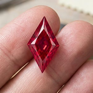 May include: A faceted, kite-shaped ruby gemstone with a deep, vibrant red color. The stone features sharp, geometric edges and a brilliant, reflective surface that catches the light, showcasing its precise cut and intense, saturated crimson hue.