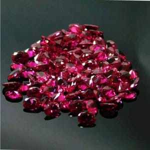 May include: A pile of faceted, deep red gemstones. The gems are scattered across a reflective black surface, catching the light and displaying multiple facets. The stones vary in size and shape, creating a sparkling, luxurious display.
